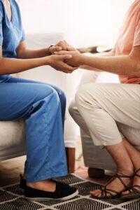 Close-up of a nurse in scrubs holding hands with a seated senior woman in a comforting gesture.